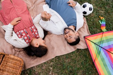 Top view of smiling asian father and daughter holding hands near football and kite in park 