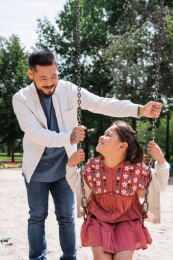 Positive asian girl sitting on swing near tattooed dad in park 