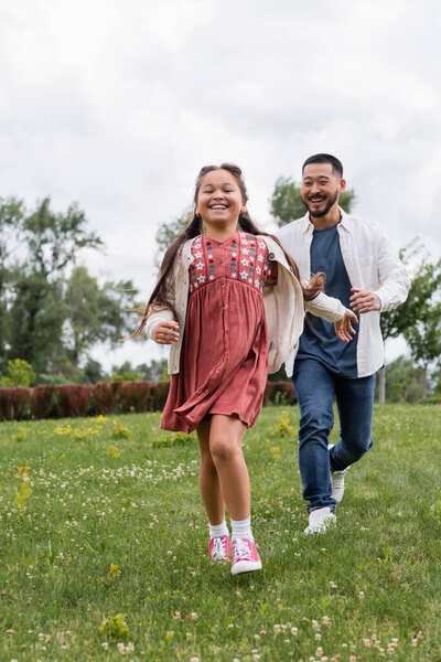 Cheerful asian girl running near father in summer park 
