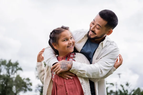 Cheerful asian father hugging preteen kid in park 