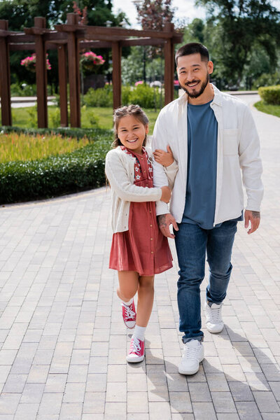 Positive asian girl holding hand of dad while walking in summer park 