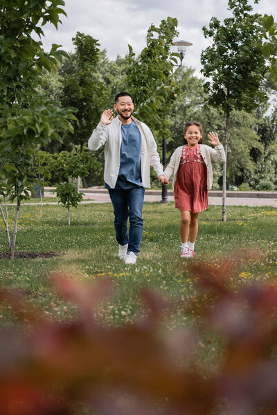 Positive asian parent and kid waving hands in summer park 