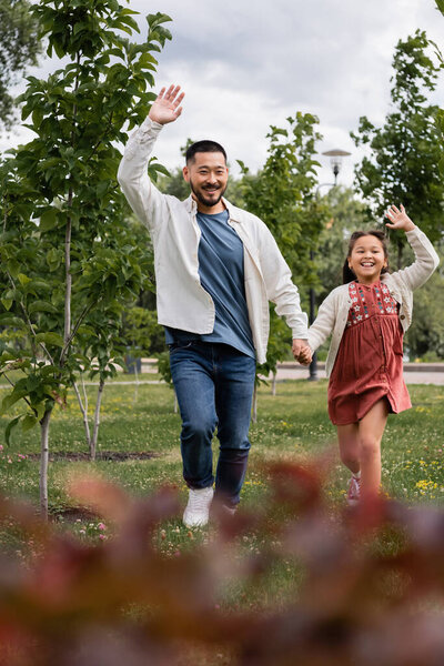 Happy asian father and child waving hands while running in park 