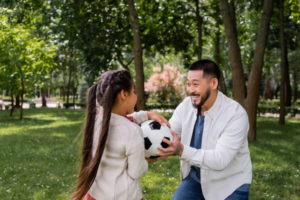 Positive asian man holding ball near daughter in park 
