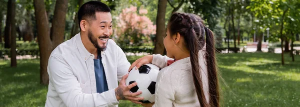 Cheerful asian man holding football near preteen daughter in park, banner 