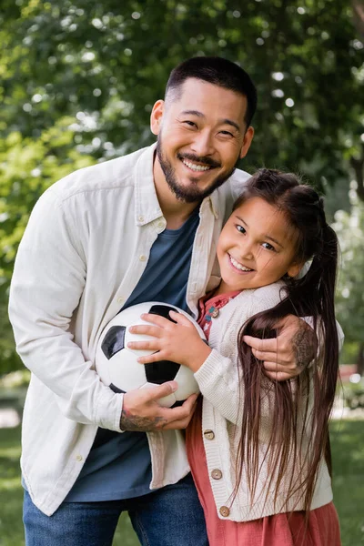 Positive asian dad and girl holding soccer ball and looking at camera in park