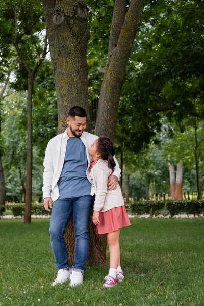 Smiling asian dad hugging and looking at daughter near tree in park