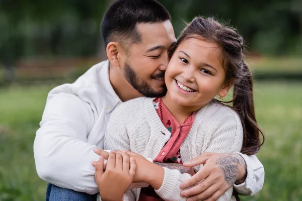 Portrait of asian dad hugging daughter looking at camera in park