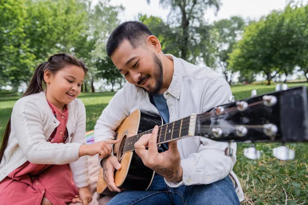 Asian father playing acoustic guitar with daughter in park