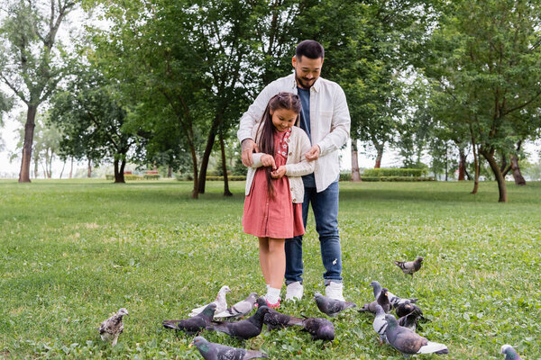 Asian father hugging daughter while feeding doves in summer park 