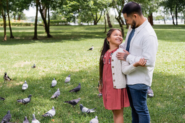 Asian man hugging preteen daughter near doves in park 