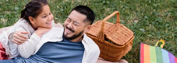 Positive asian kid looking at dad near flying kite and wicker basket in park, banner