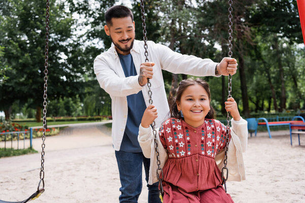Smiling father standing near asian daughter on swing in park 