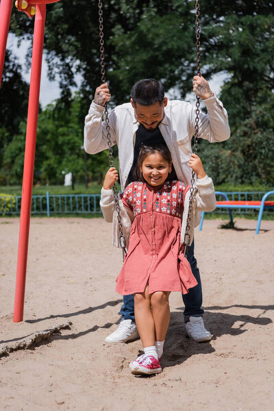 Positive preteen girl sitting on swing near asian dad in park 
