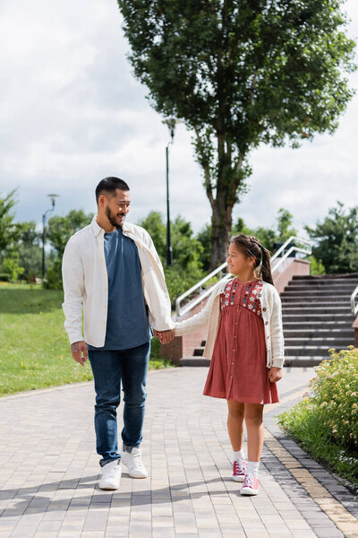 Asian parent and kid holding hands while walking in summer park 