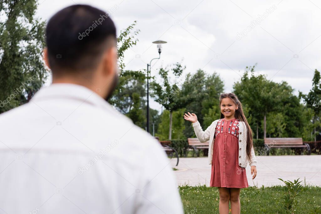 Smiling asian girl waving hand at blurred dad in summer park 
