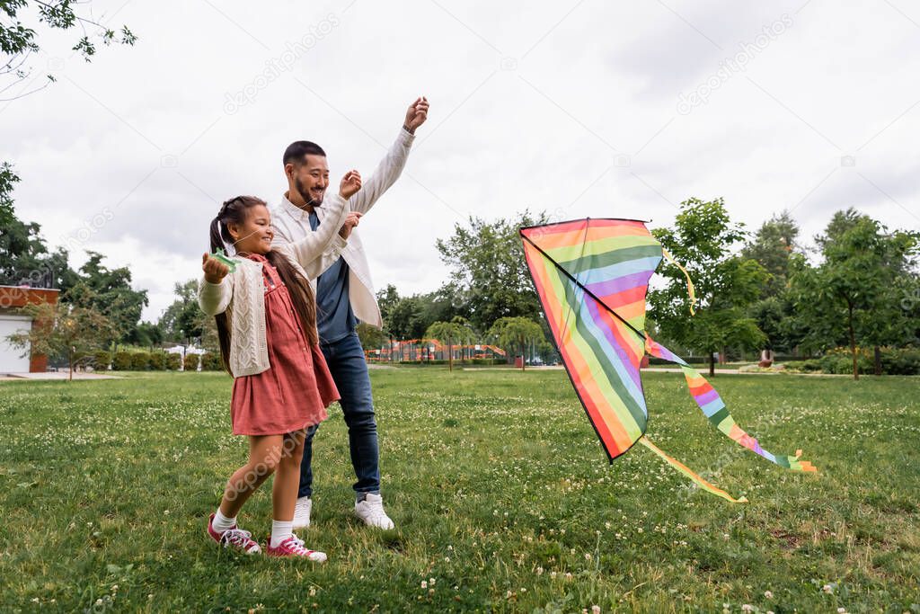 Smiling asian parent and kid looking at flying kite on lawn in park 