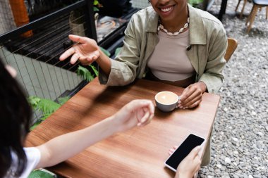Cropped view of african american woman talking to friend with smartphone near coffee in outdoor cafe 