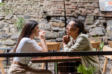 Side view of cheerful multiethnic friends talking on terrace of cafe 