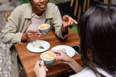 Blurred woman holding cup of cappuccino near african american friend in outdoor cafe 