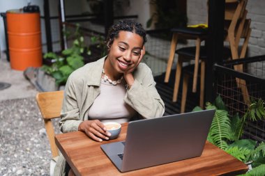 Smiling african american woman holding cup of coffee and looking at laptop in outdoor cafe 