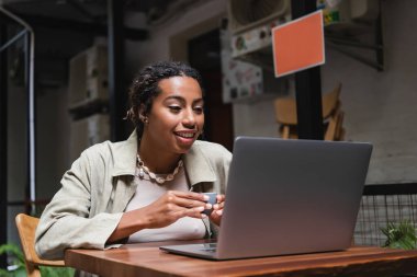 African american woman holding cup and looking at laptop on terrace of cafe 
