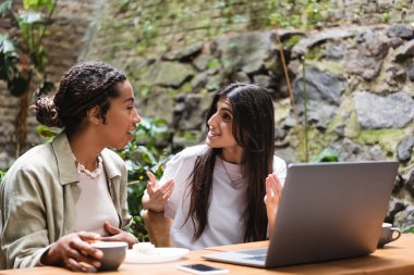 Interracial girlfriends talking near gadgets and coffee in outdoor cafe 