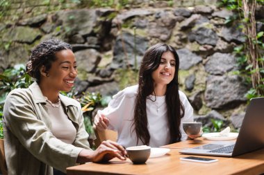 Smiling interracial girlfriends with cups of coffee looking at laptop in outdoor cafe  