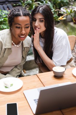 Woman telling secret to african american friend near devices and coffee in outdoor cafe 