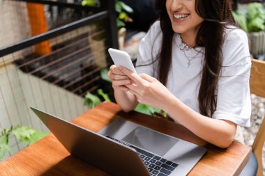 Cropped view of smiling woman using smartphone near laptop in outdoor cafe 