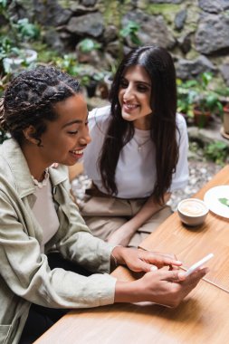 High angle view of smiling african american woman using mobile phone near friend and coffee in outdoor cafe 