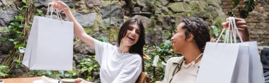 Cheerful interracial girlfriends holding shopping bags in outdoor cafe, banner 