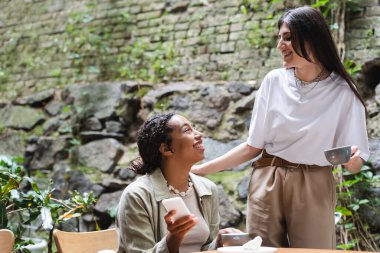 Side view of smiling interracial friends with coffee and smartphone looking at each other in outdoor cafe 