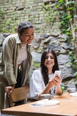 Smiling woman holding smartphone near african american friend and cups of coffee in outdoor cafe 