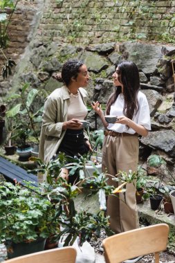 Side view of smiling african american woman holding cup and talking to friend in outdoor cafe 