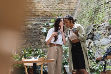 Woman holding cup and telling secret to african american girlfriend in outdoor cafe 