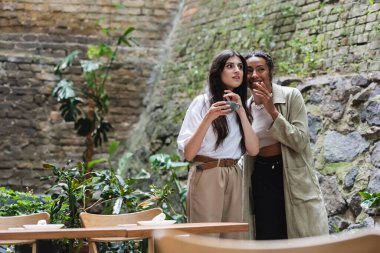 African american woman telling secret to amazed friend with coffee on terrace of cafe 