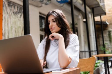 Brunette woman using laptop near blurred cellphone on terrace of cafe 