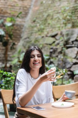 Happy woman holding coffee and dessert and smiling at camera near smartphone on terrace of cafe 