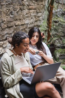 Cheerful african american woman using laptop near friends with cup of coffee in outdoor cafe 