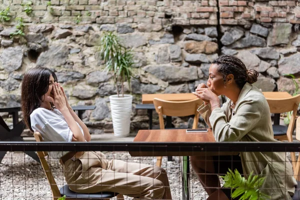 Side view of woman laughing near african american friend with coffee in outdoor cafe