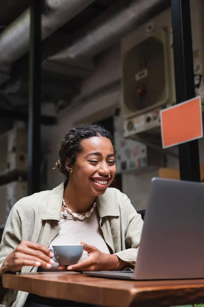 Позитивна Афро Американська Жінка Тримає Чашку Кави Дивиться Ноутбук Терасі — стокове фото
