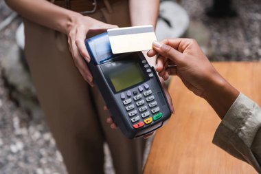 Cropped view of waitress holding payment terminal near african american client with credit card on cafe terrace 