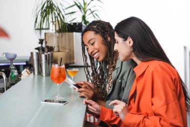 Cheerful multiethnic girlfriends using smartphone near cocktails in cafe 
