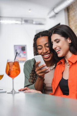 Smiling african american woman using smartphone near friend and blurred cocktails in cafe 