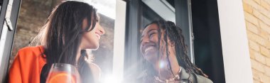 Low angle view of smiling multiethnic girlfriends talking near cocktail in cafe, banner 