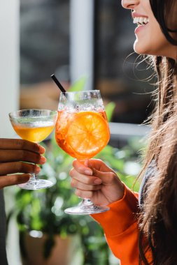 Cropped view of cheerful woman toasting cocktail with african american friend in cafe  
