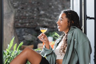 Side view of cheerful african american woman holding cocktail near window in cafe 
