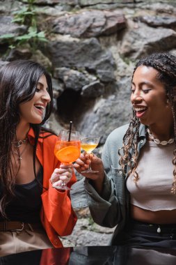 Positive multiethnic girlfriends clinking with cocktails on terrace of cafe 
