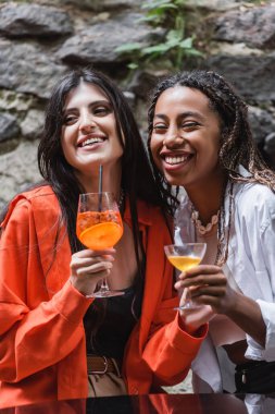 Smiling african american woman holding cocktail near girlfriend on terrace of cafe 